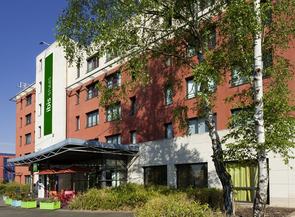 Un edificio moderno con un letrero vertical verde, varias ventanas y un área de acceso cubierta con asientos al aire libre.