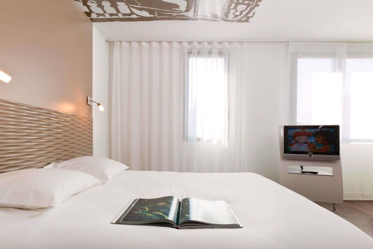 Quarto de hotel com cama, televisão de tela plana, um livro e janela com cortinas finas.