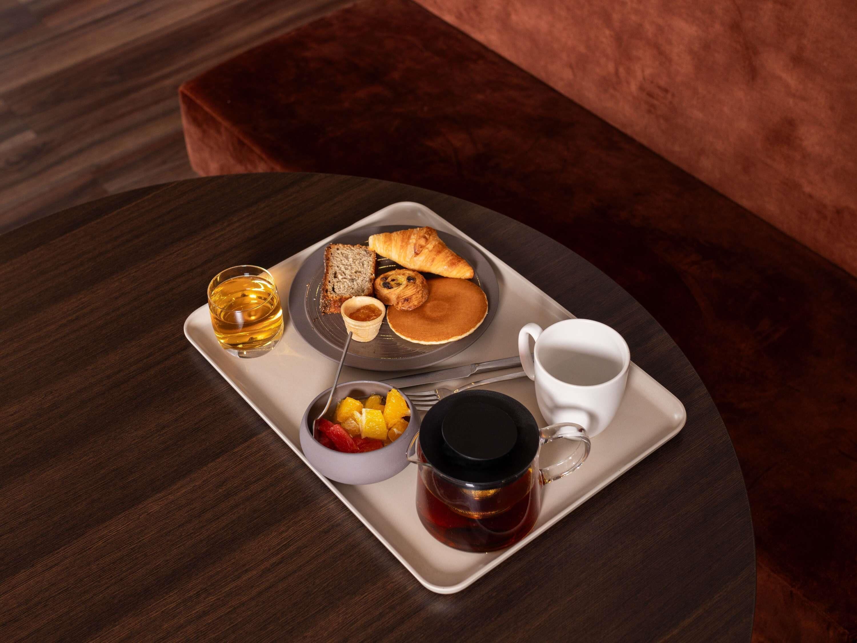 Una bandeja con alimentos para el desayuno, como tostadas, croissants, panqueques, una taza, té y un tazón de fruta.