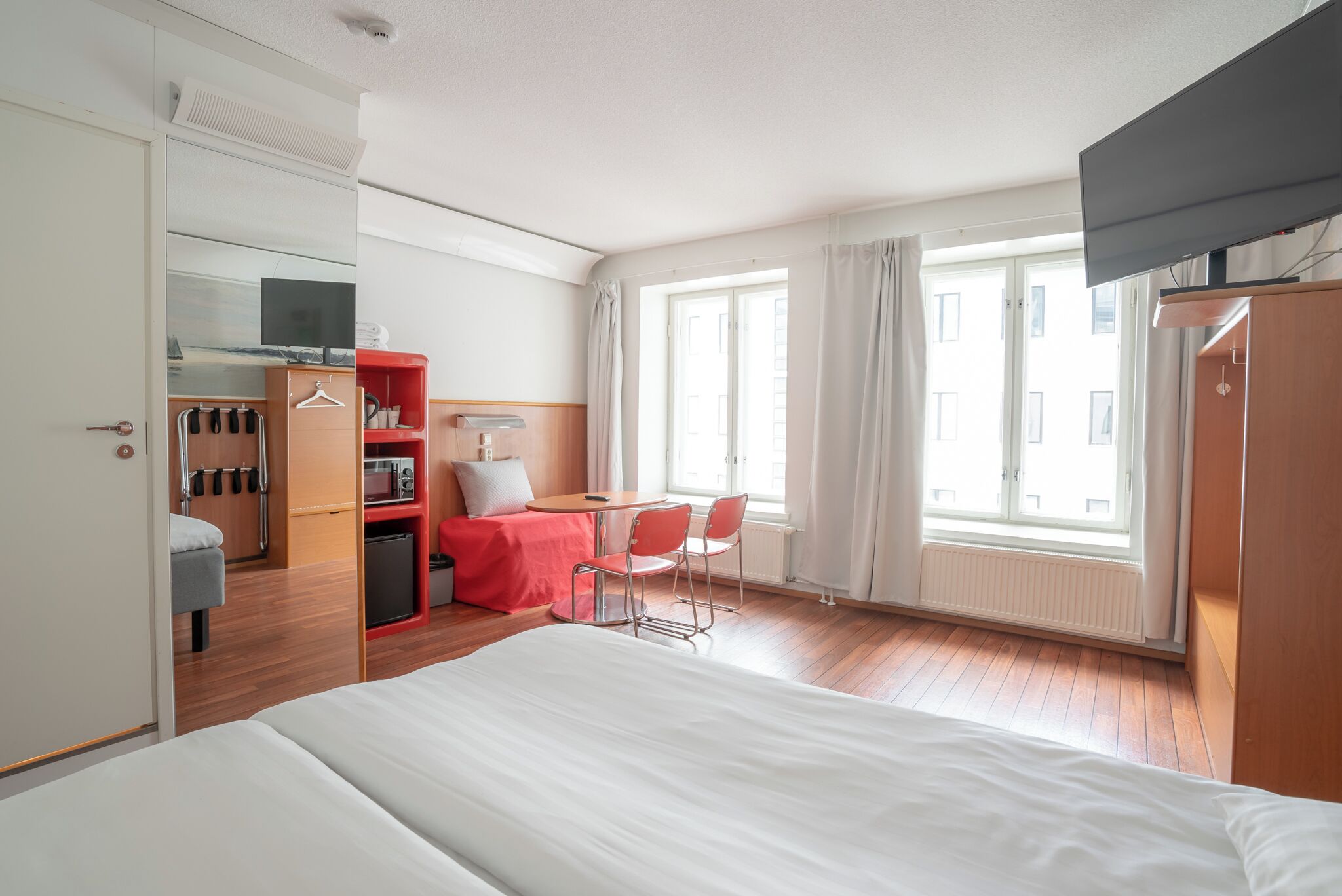 Ein Hotelzimmer mit einem großen Bett, einem Schreibtisch mit Stuhl, einem Kleiderschrank, einem Spiegel und einem Fenster mit Vorhängen.