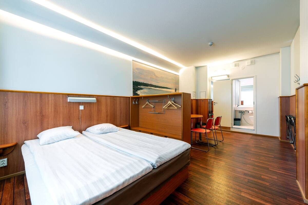 A hotel room with a wooden headboard, a bed with white linens, a wooden floor, a seating area with chairs, and a bathroom visible through an open door.
