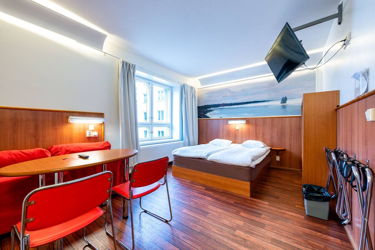 A hotel room with a bed, a desk with a chair, a TV mounted on the wall, and a window with curtains.