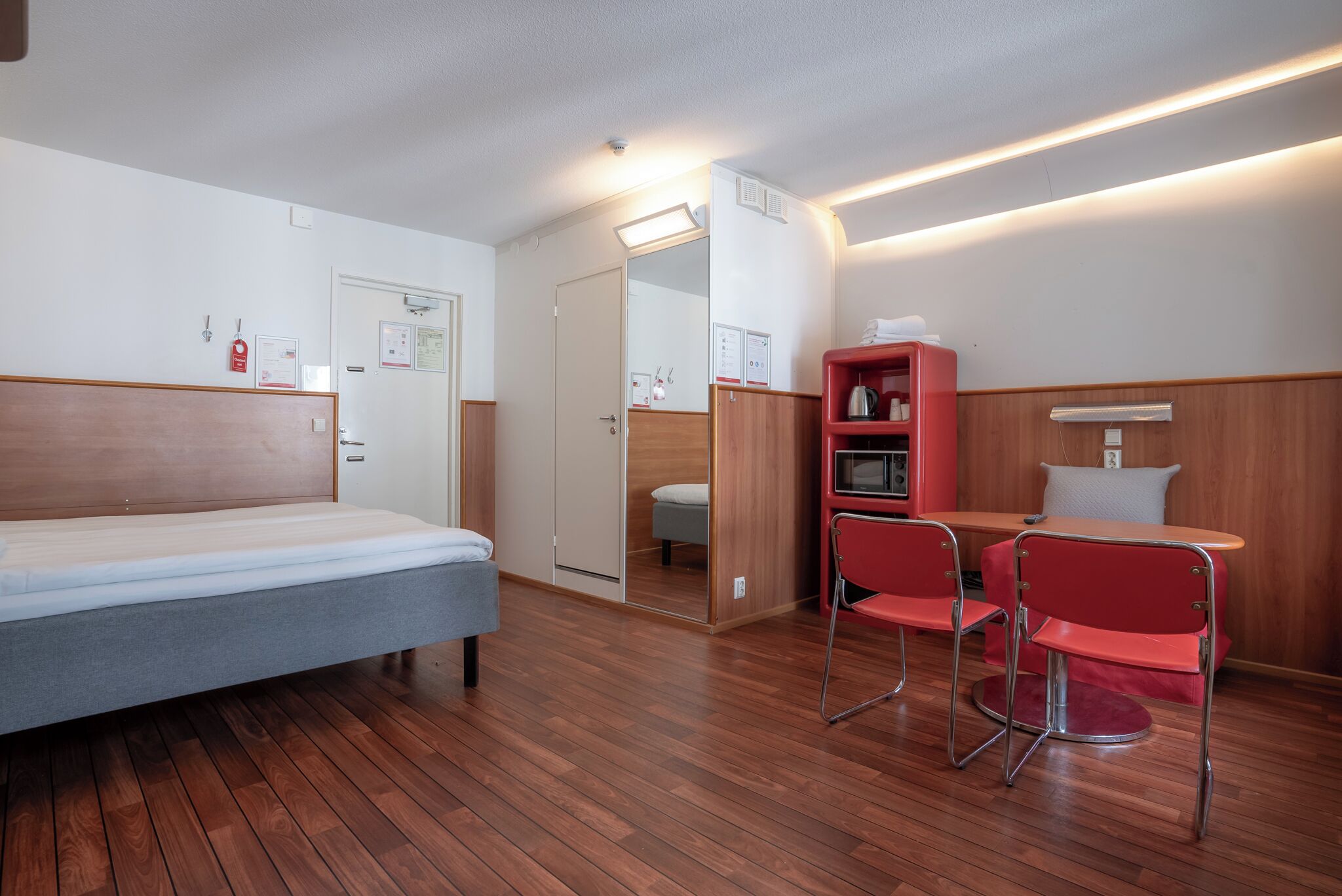A hotel room with a bed, a small table with two red chairs, a red cabinet, and a mirror.