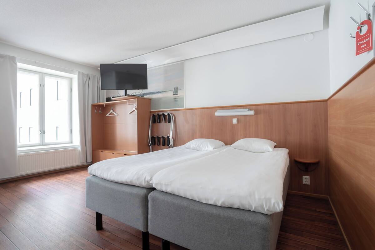 A double bed with grey upholstered headboards, a wardrobe with a mirror, a television mounted above the bed, and a window with curtains.