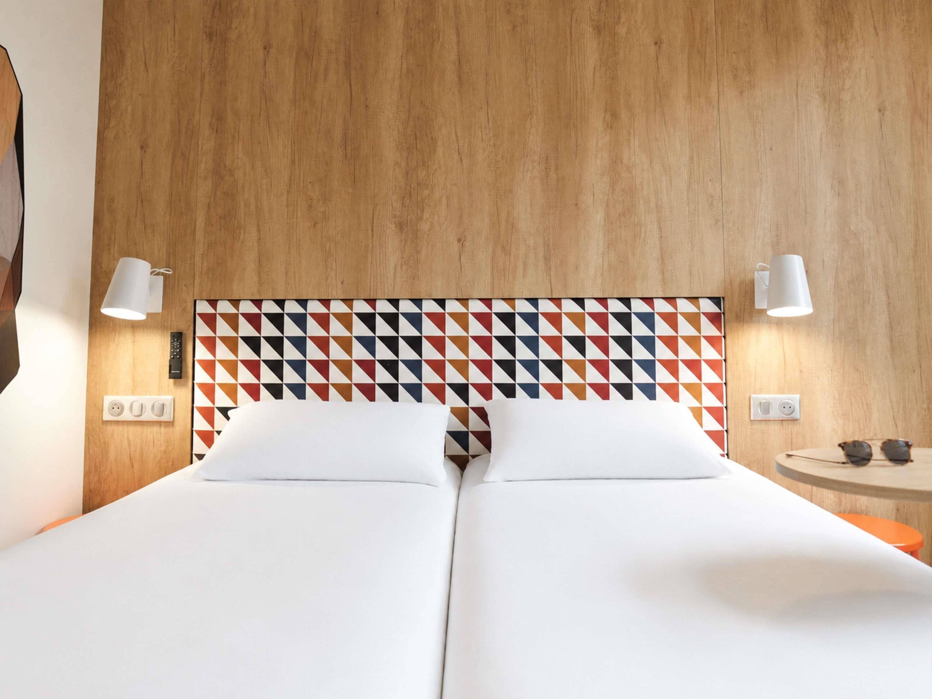 Una cama doble con cabecera de diseño geométrico, lámparas de noche, una mesita con gafas de sol y un enchufe.