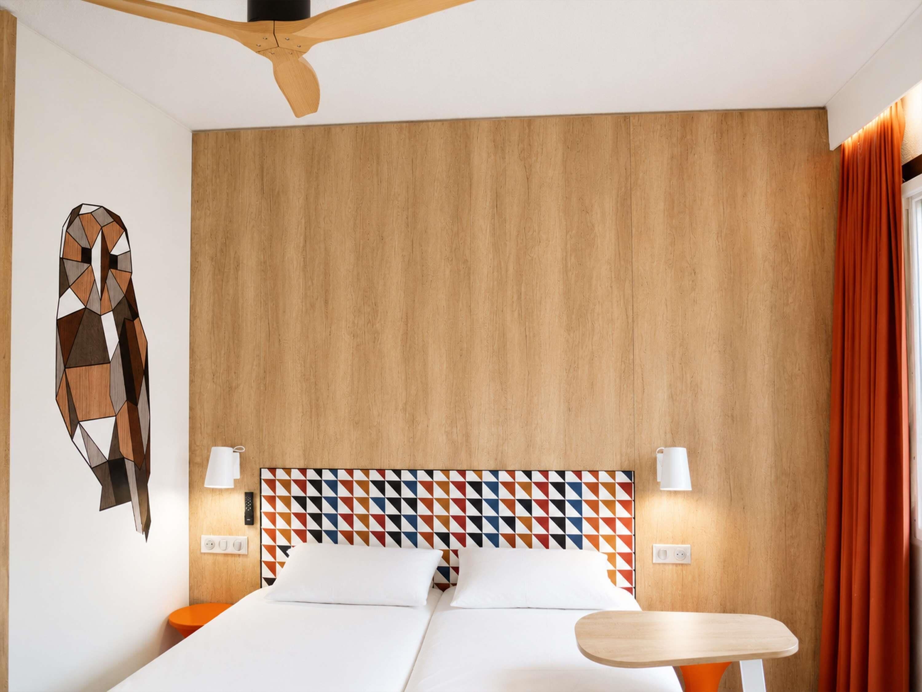 A hotel room with a wooden headboard, a geometric-patterned wall, a bed with white linens, two bedside lamps, and a ceiling fan.