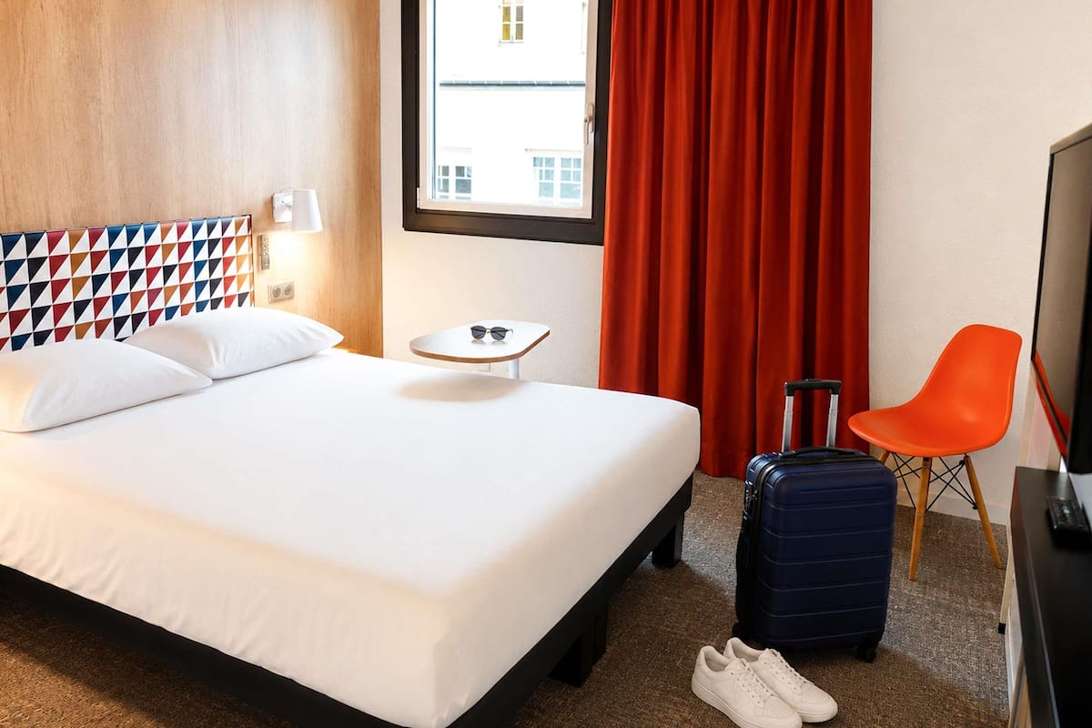 A hotel room with a bed, a small table, a chair, a suitcase, and a TV.