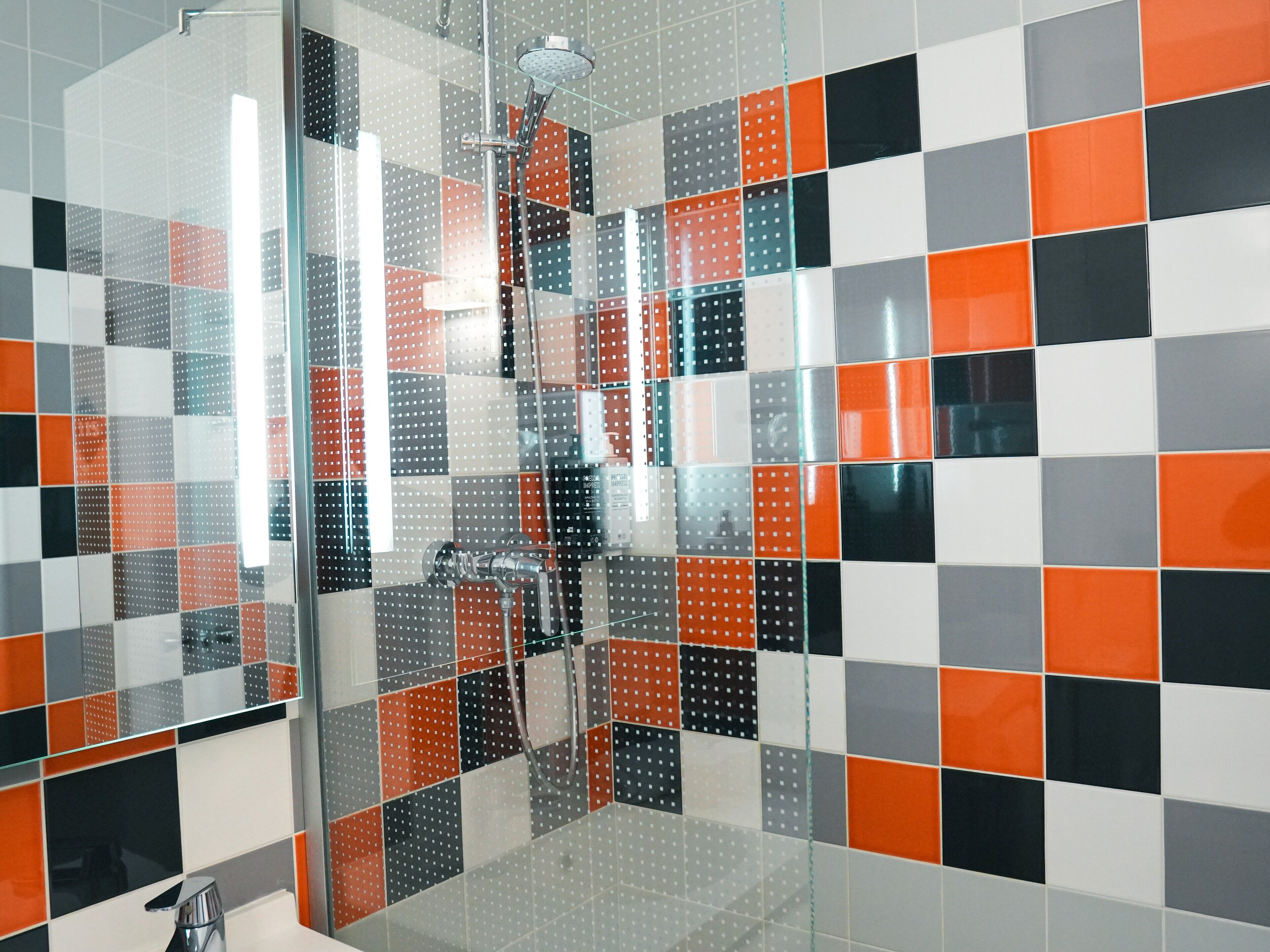 Un baño con cabina de ducha de vidrio, paredes cubiertas de azulejos en blanco, negro y naranja, y un cabezal de ducha visible.