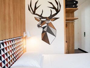 A hotel room with a geometric-patterned headboard, a wall-mounted deer head artwork, a bedside lamp, and a wooden wardrobe.