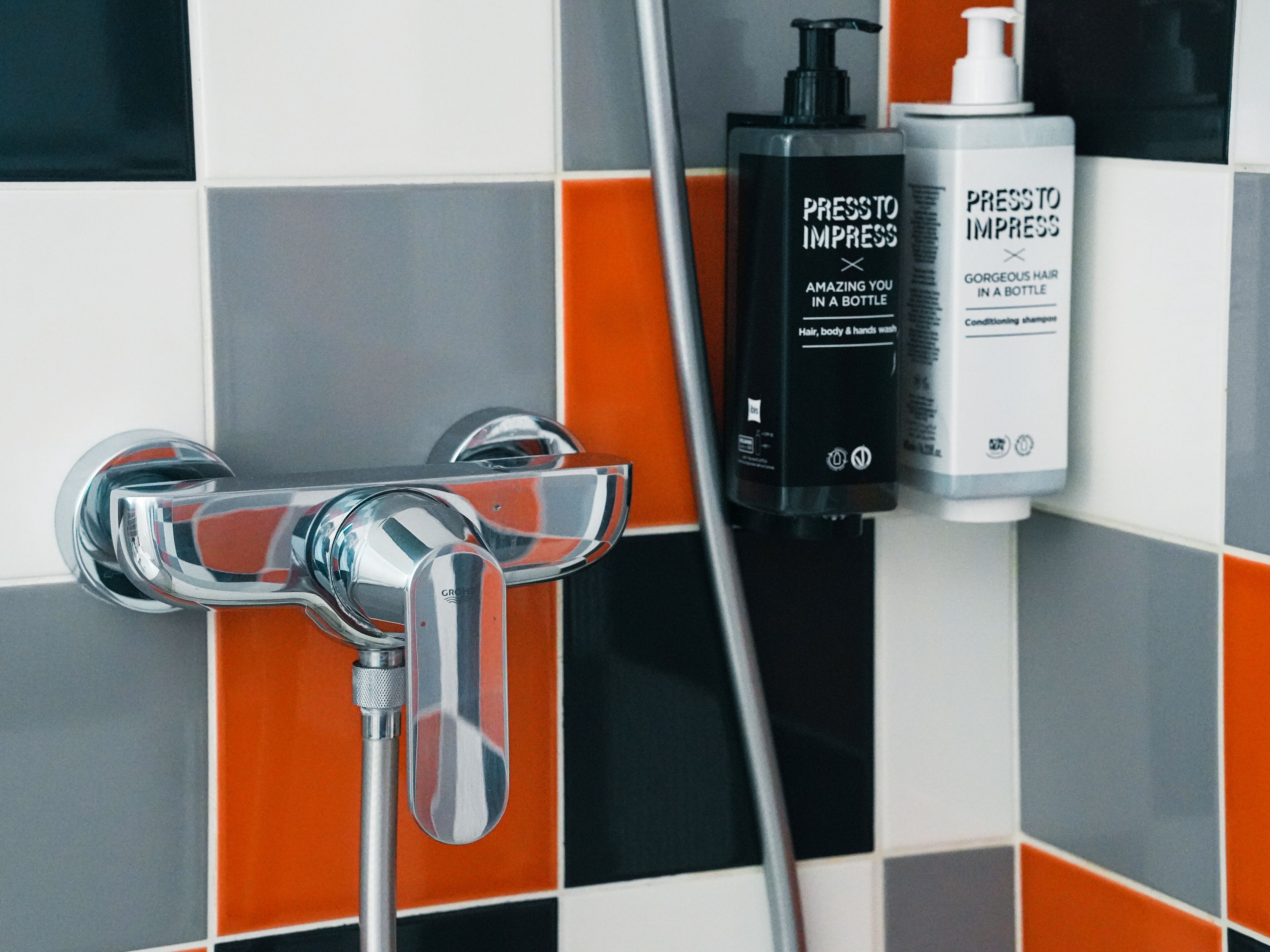 Un baño con un grifo cromado, cabezal de ducha y dos botellas de champú y acondicionador 'Press to Impress'.