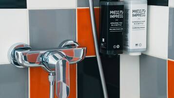 Un baño con un grifo cromado, cabezal de ducha y dos botellas de champú y acondicionador 'Press to Impress'.