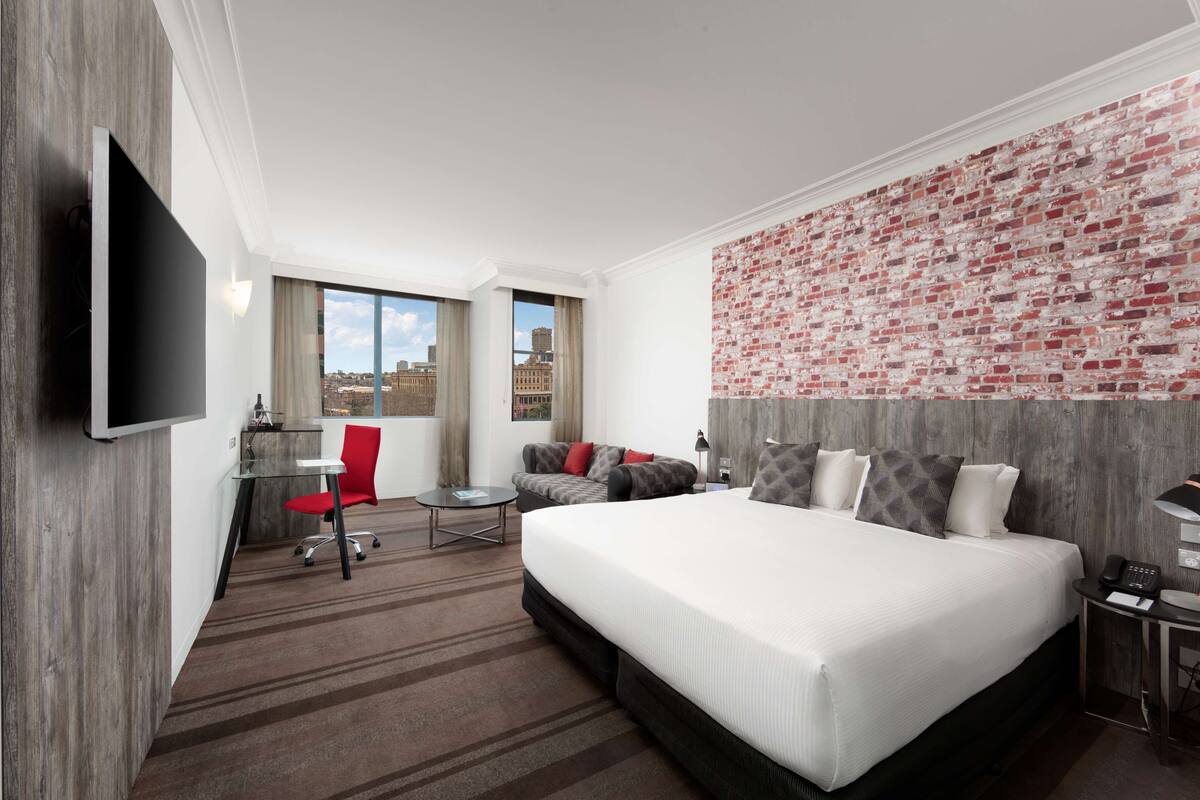A hotel room with a large bed, a desk with a chair, a TV, and a brick-patterned wall.