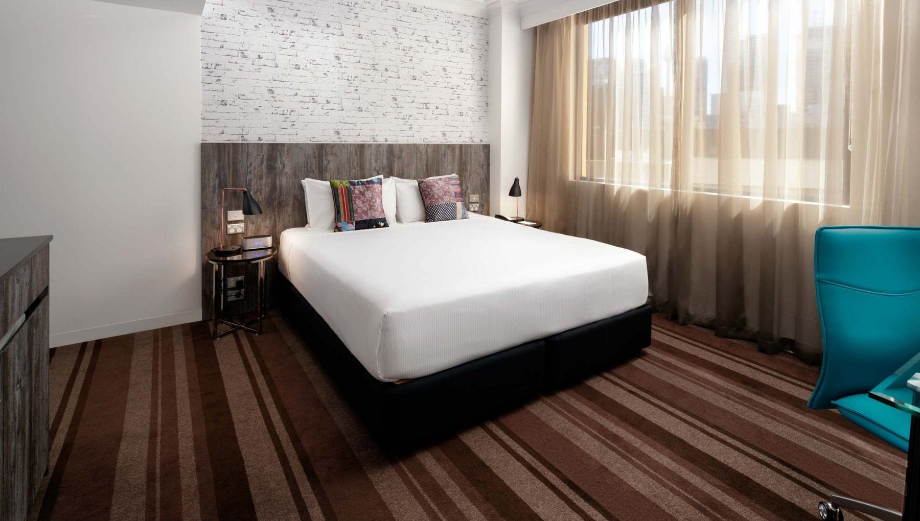 Een hotelkamer met een bed, nachtkastje, lamp, stoel en raam met gordijnen.
