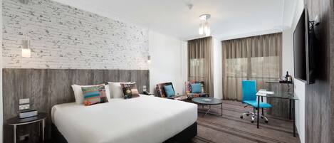 A hotel room with a large bed, a desk, two chairs, a TV, and a brick-patterned wall.