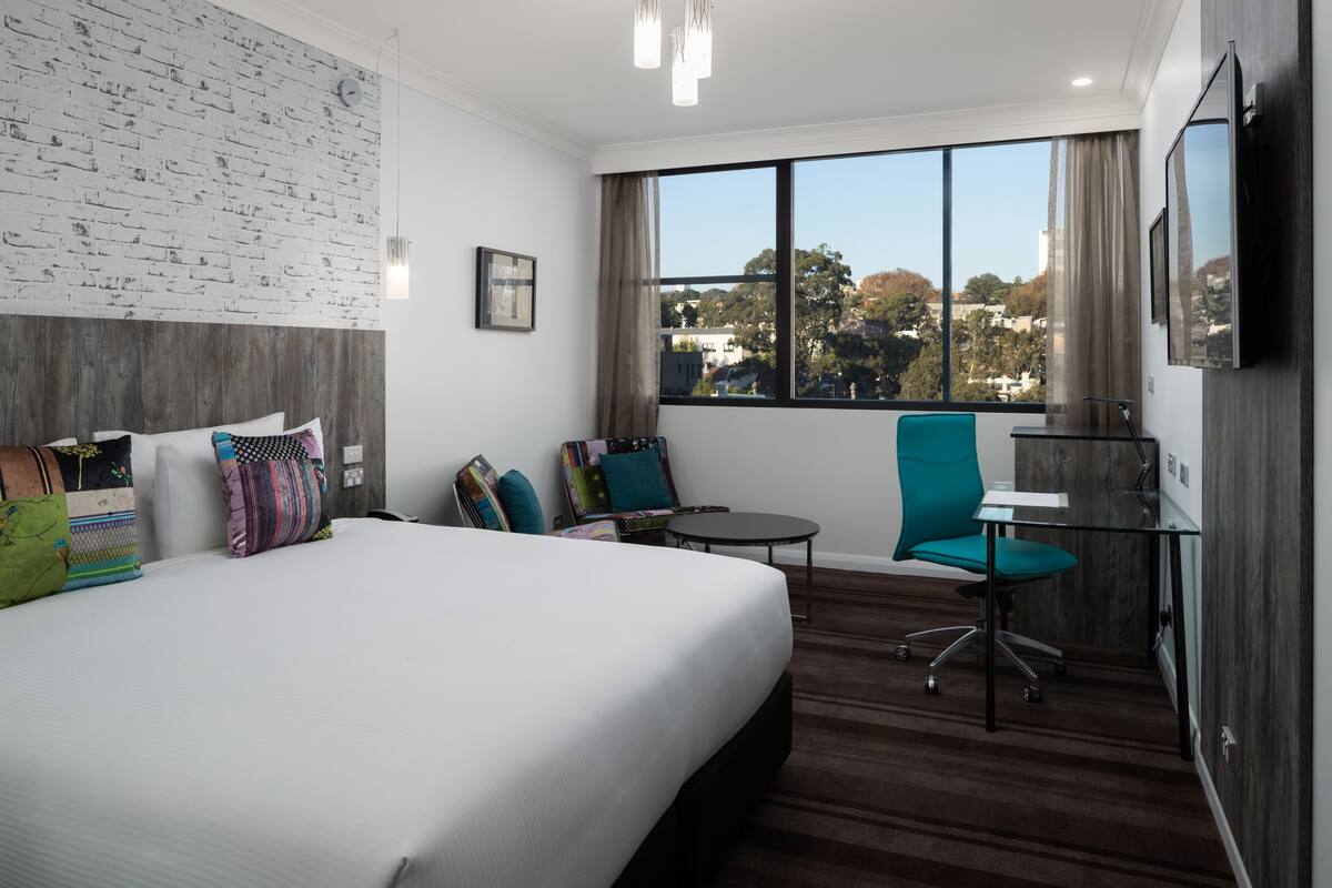 A hotel room with a large bed, a desk, a chair, and a view of the city.