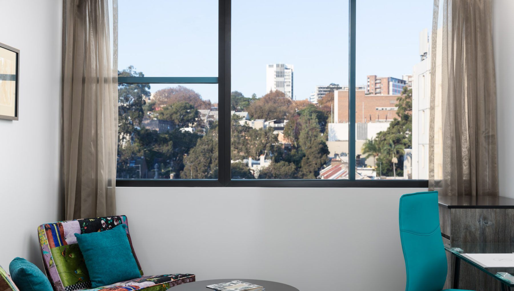 A room with a large window, a teal chair, a small table, and a colorful cushion.