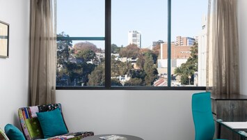 Una habitación con un ventanal grande, una silla color turquesa, una mesita y un cojín colorido.
