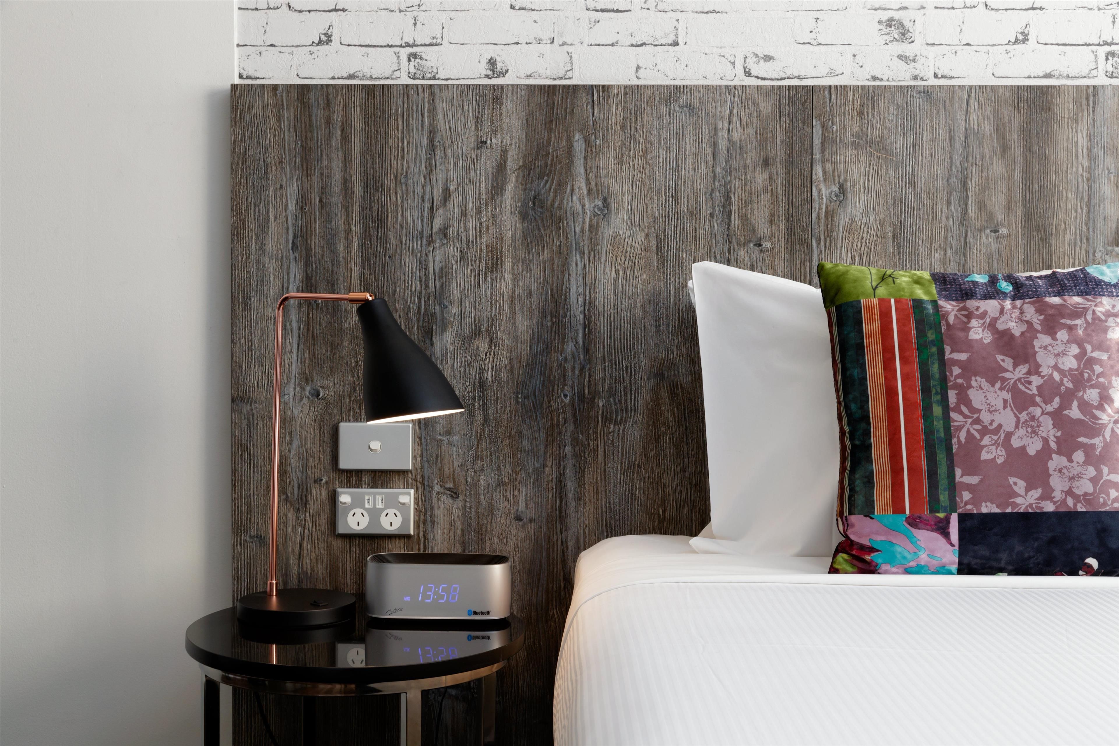 A neatly made bed with a white pillow and a colorful pillow, a round bedside table with a lamp, a clock, and a power outlet.