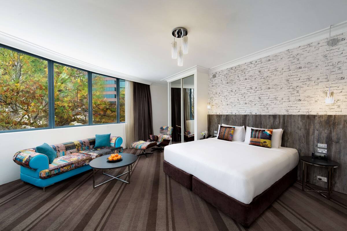 A modern hotel room with a large bed, a sofa with colorful cushions, a small round table with fruit, and a view of autumn trees through large windows.