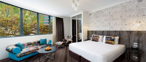 Une chambre d’hôtel moderne avec un grand lit, un canapé agrémenté de coussins colorés, une petite table ronde avec des fruits, et une vue sur les arbres d’automne à travers de grandes fenêtres.