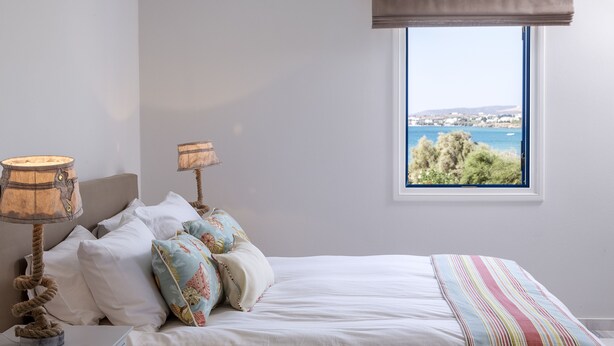 A bedroom with a bed, two lamps, a window with a view of the sea, and a striped blanket. - Poseidon of Paros (Paros)