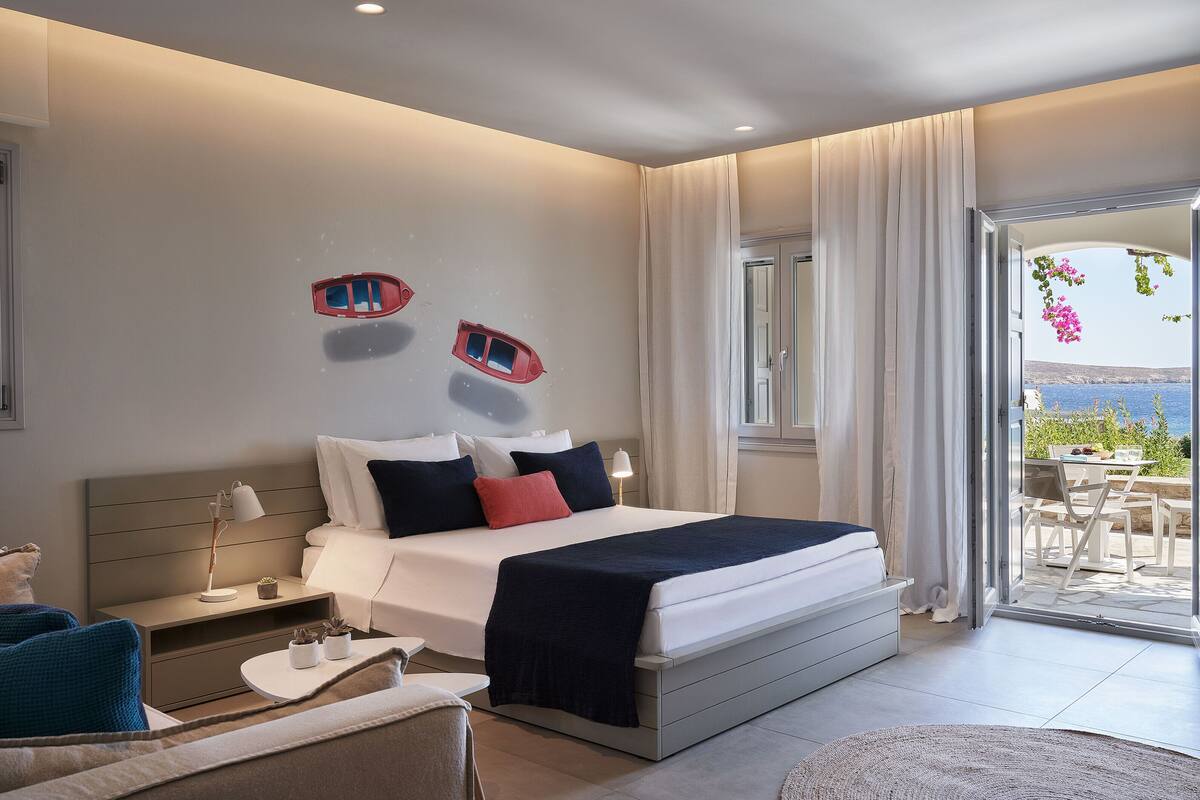 Ein modernes Schlafzimmer mit einem großen Bett, einem Nachttisch, einer kleinen Anrichte und Meerblick durch eine Schiebetür.