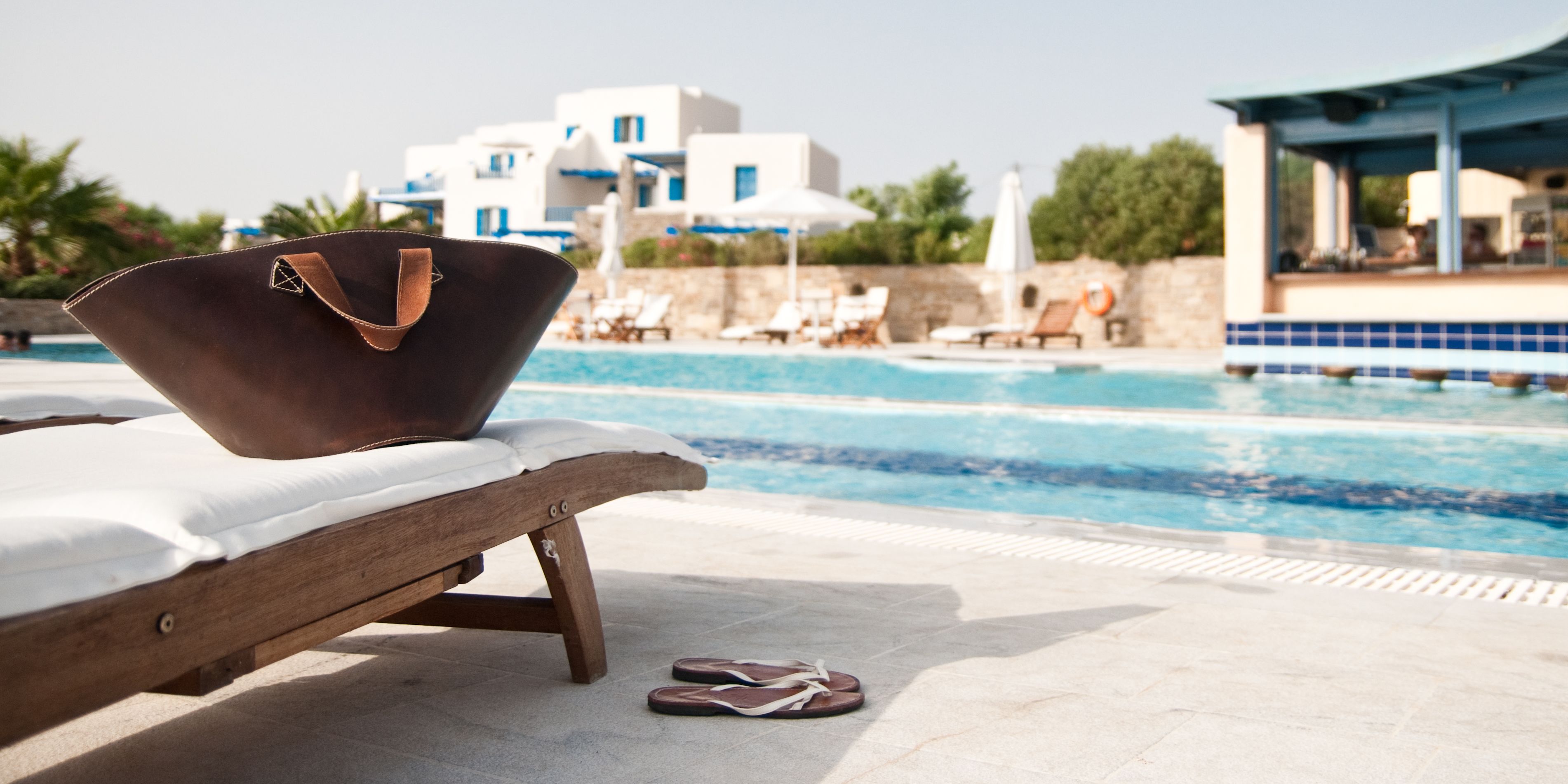 A poolside lounge with a wooden bench, a brown bag, and a pair of flip-flops. In the background, there are white buildings with blue accents and a clear blue pool.