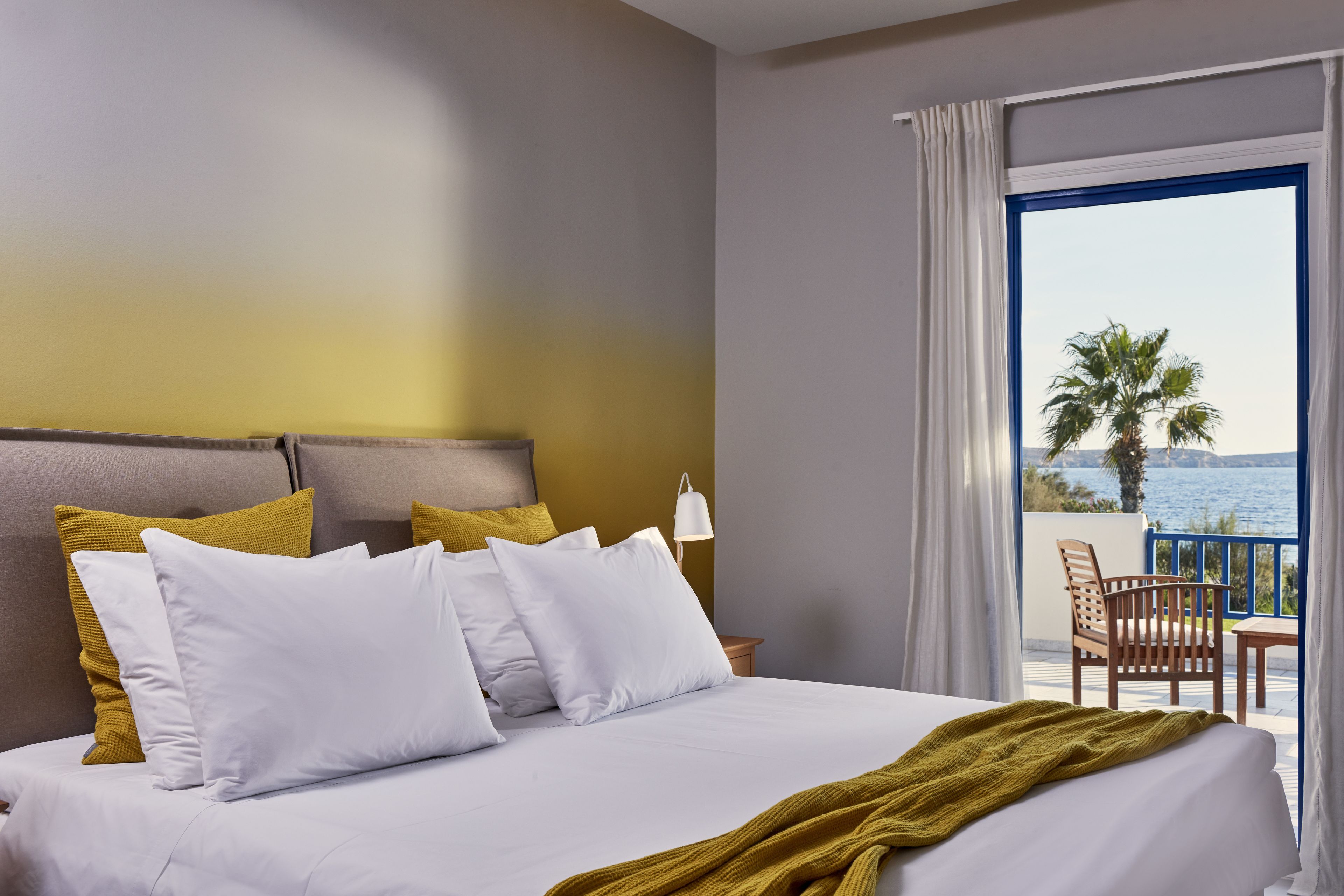 A hotel room with a bed, white and mustard pillows, a wooden headboard, a nightstand with a lamp, and a balcony with a view of the sea and palm trees.
