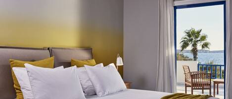 A hotel room with a bed, white and mustard pillows, a wooden headboard, a nightstand with a lamp, and a balcony with a view of the sea and palm trees.