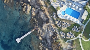 An aerial view of a resort with a swimming pool, lounge area, and rocky coastline.