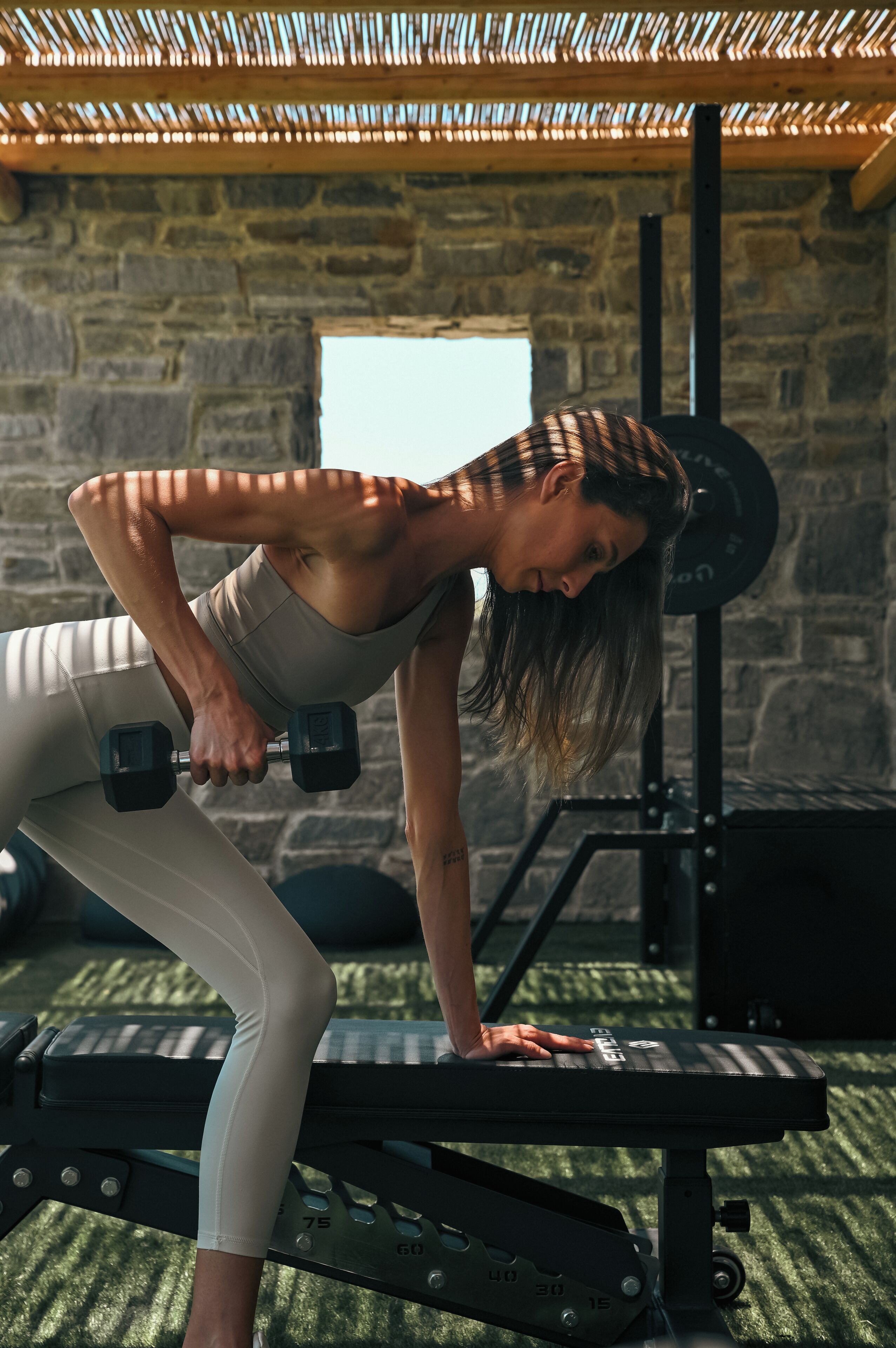 A person performing a workout on a bench with dumbbells, in a room with stone walls and a thatched roof.