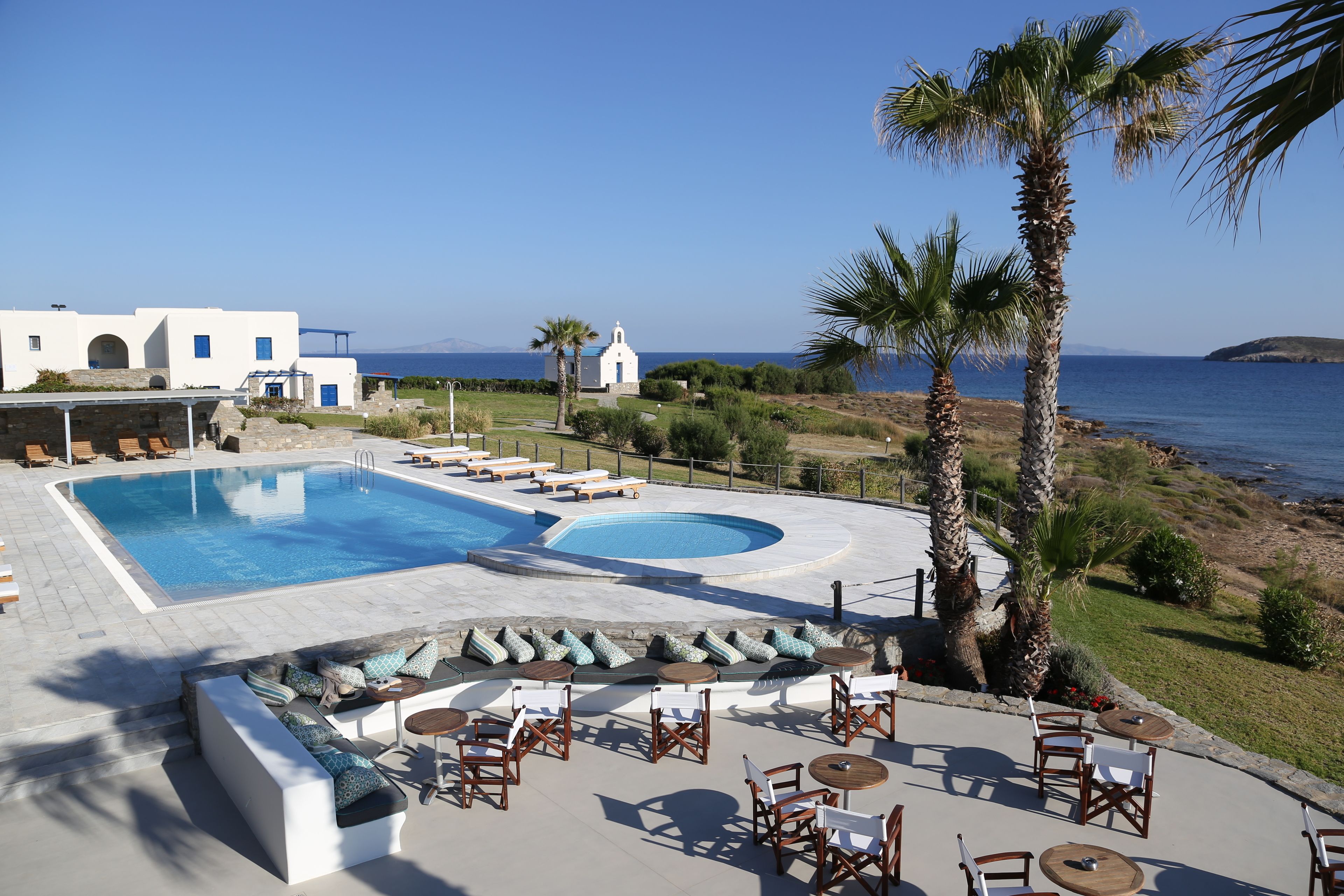 Un espace piscine avec des chaises longues et des tables, un bâtiment blanc agrémenté de touches de bleu, et une vue sur la mer.