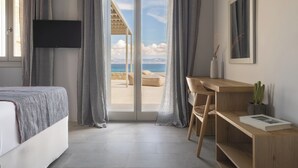 A bedroom with a bed, a desk, a chair, a TV, and a view of the sea through a sliding door.