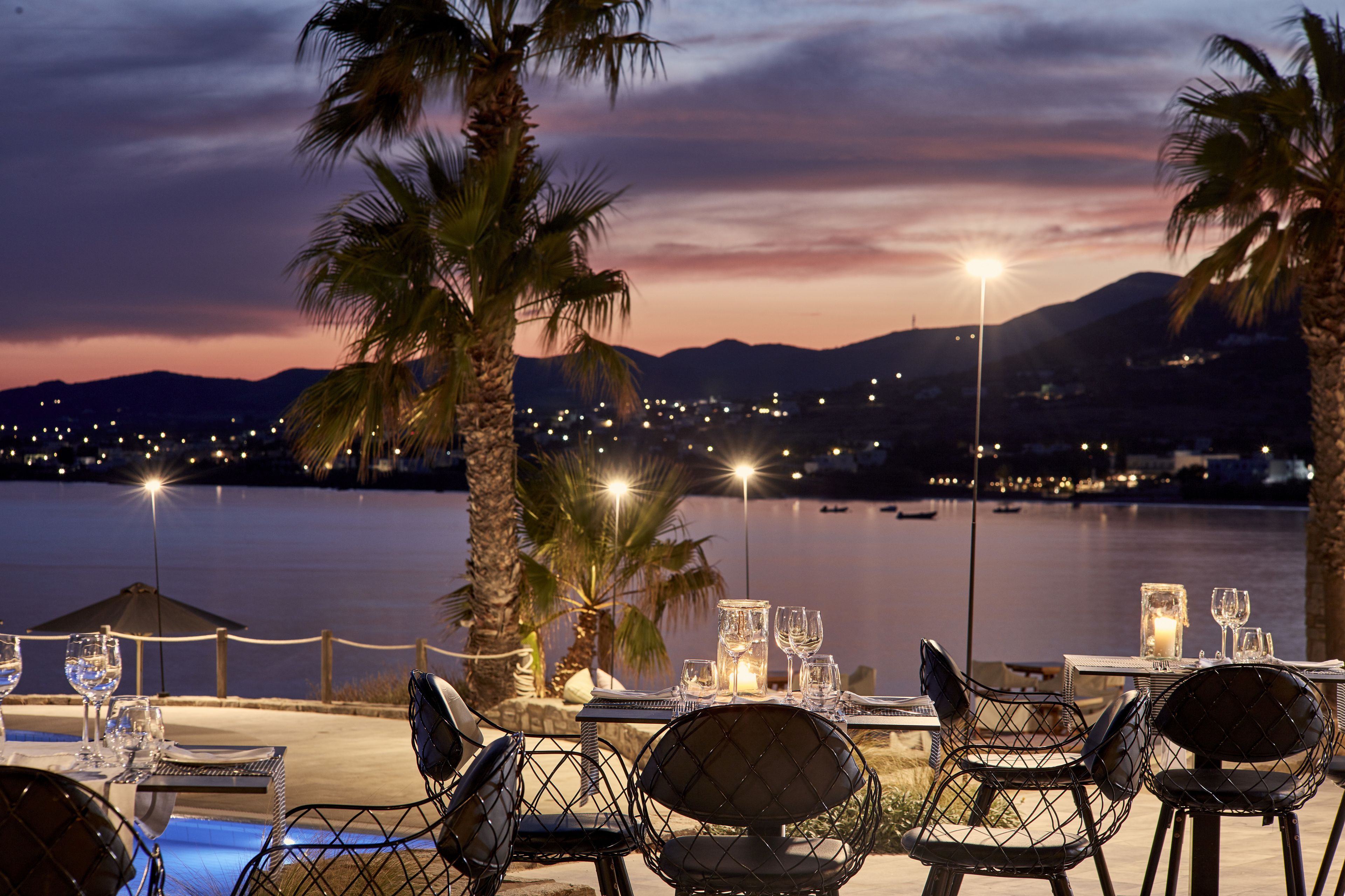 Un área de comedor al aire libre con mesas y sillas, con vista a un cuerpo de agua y luces de la ciudad al fondo.