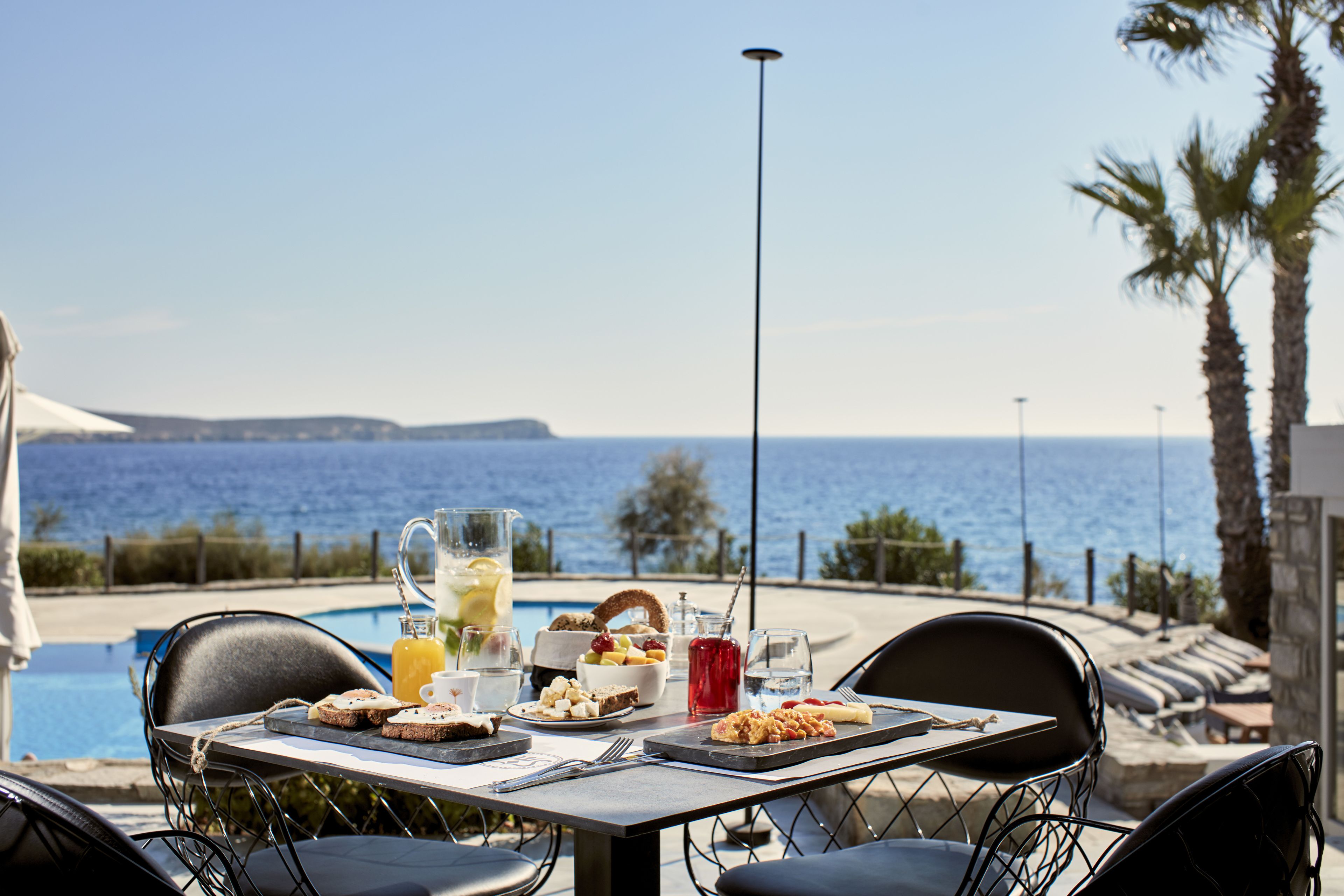 Ein Esstisch mit Frühstücksangeboten und Getränken, mit Blick auf einen Pool und das Meer.
