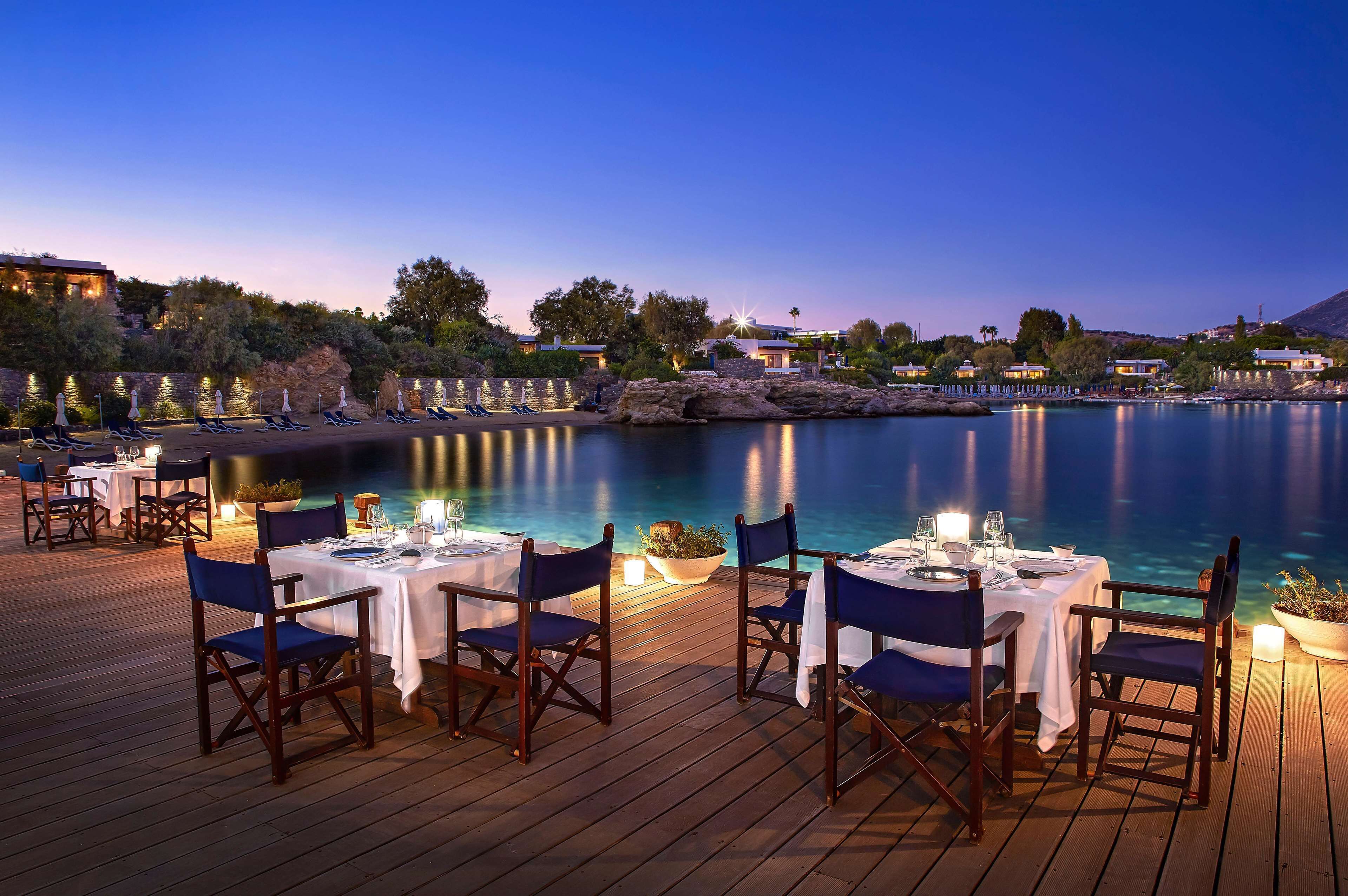 Un espace de restauration situé au bord de l’eau, avec des tables et des chaises, offrant une vue sur une étendue d’eau calme et un paysage montagneux en arrière-plan.