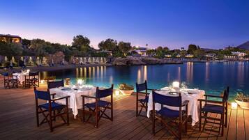 Un espace de restauration situé au bord de l’eau, avec des tables et des chaises, offrant une vue sur une étendue d’eau calme et un paysage montagneux en arrière-plan.
