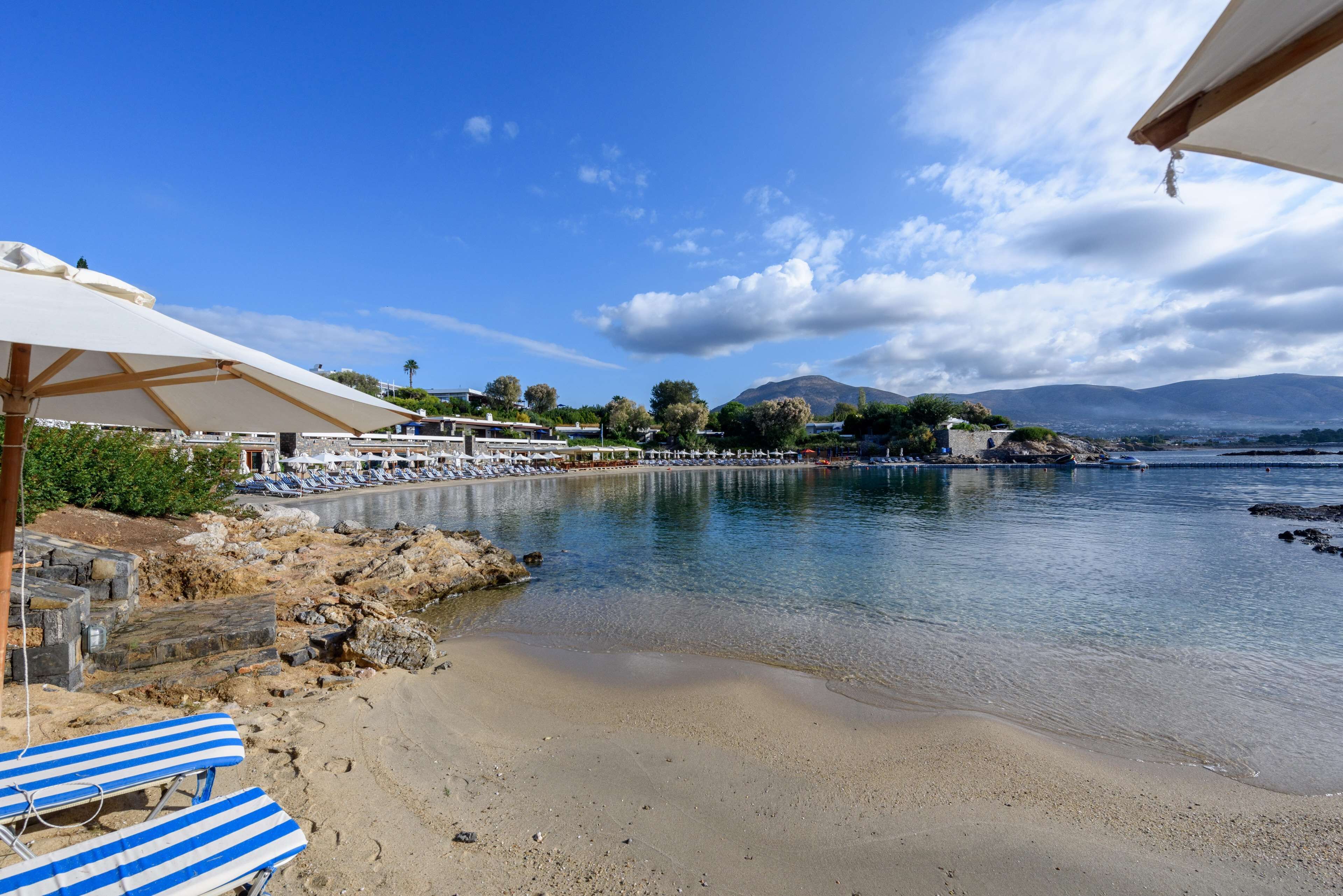Una playa con tumbonas y sombrillas, un litoral rocoso y vistas al mar y montañas distantes.