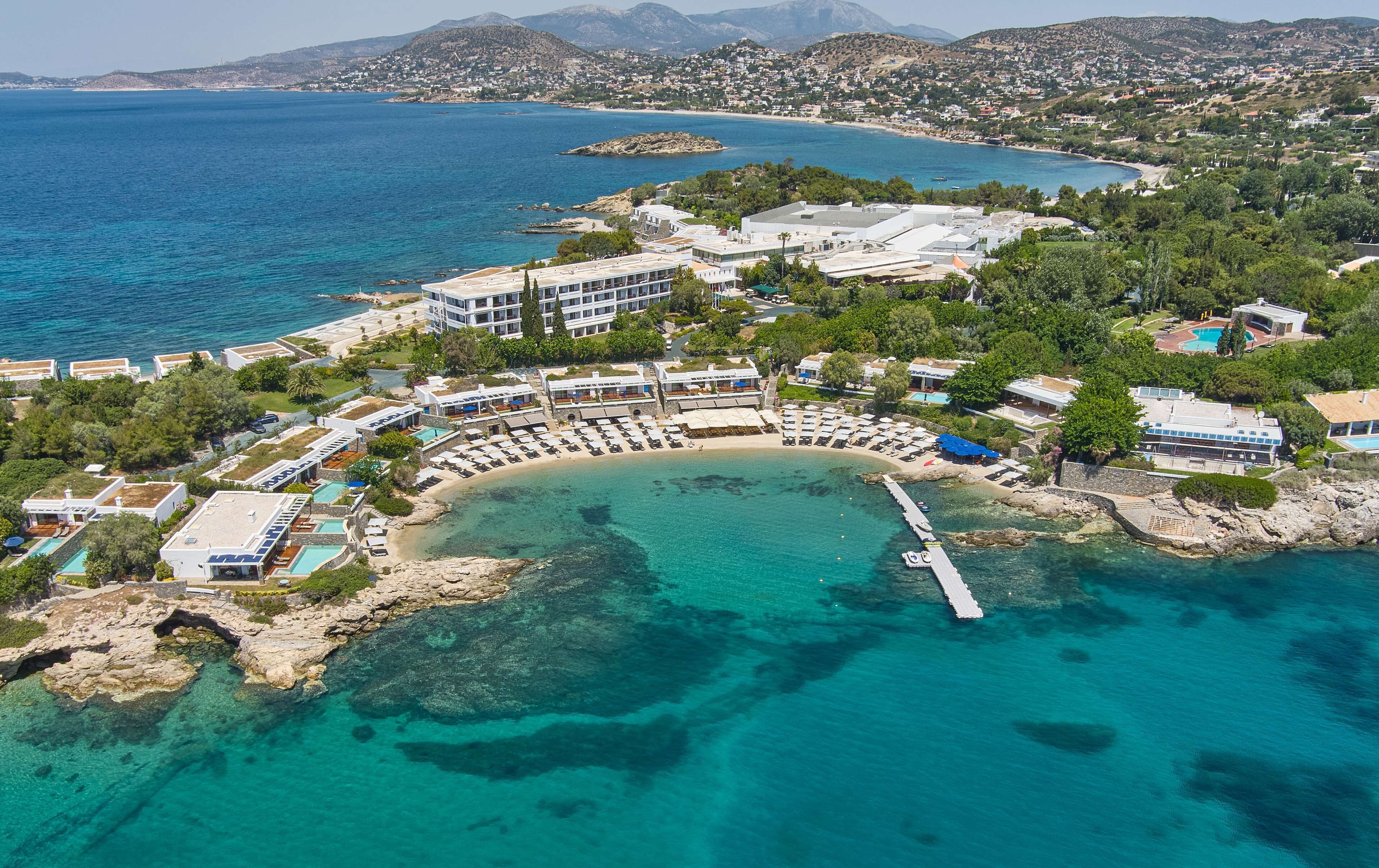 An aerial view of a coastal resort with a marina, beach, and buildings with balconies overlooking the sea.