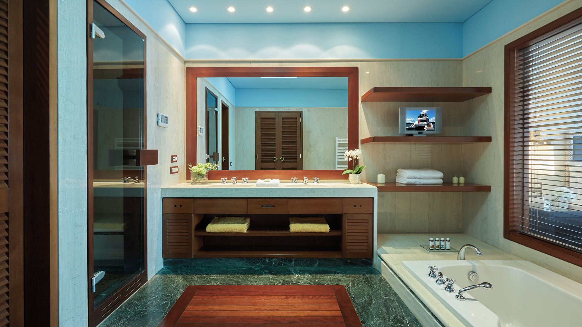 Ein modernes Badezimmer mit großem Spiegel, zwei Waschbecken, einer Badewanne und einem Fernseher über dem Spiegel.
