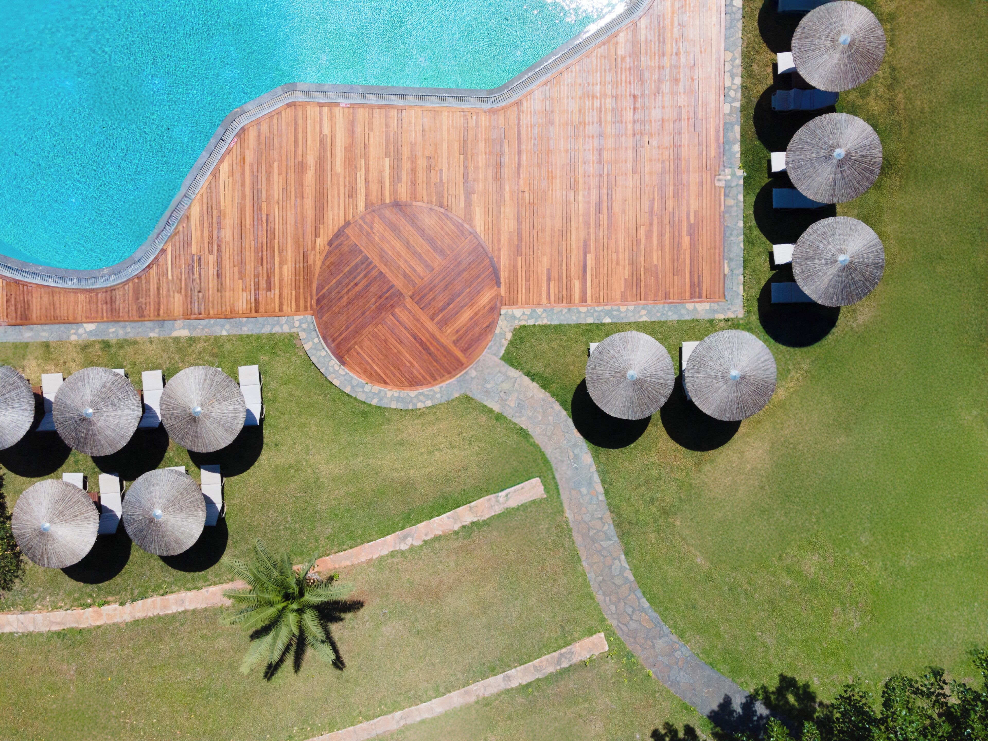 An aerial view of a resort with a swimming pool, wooden deck, lounge chairs, umbrellas, and a palm tree.