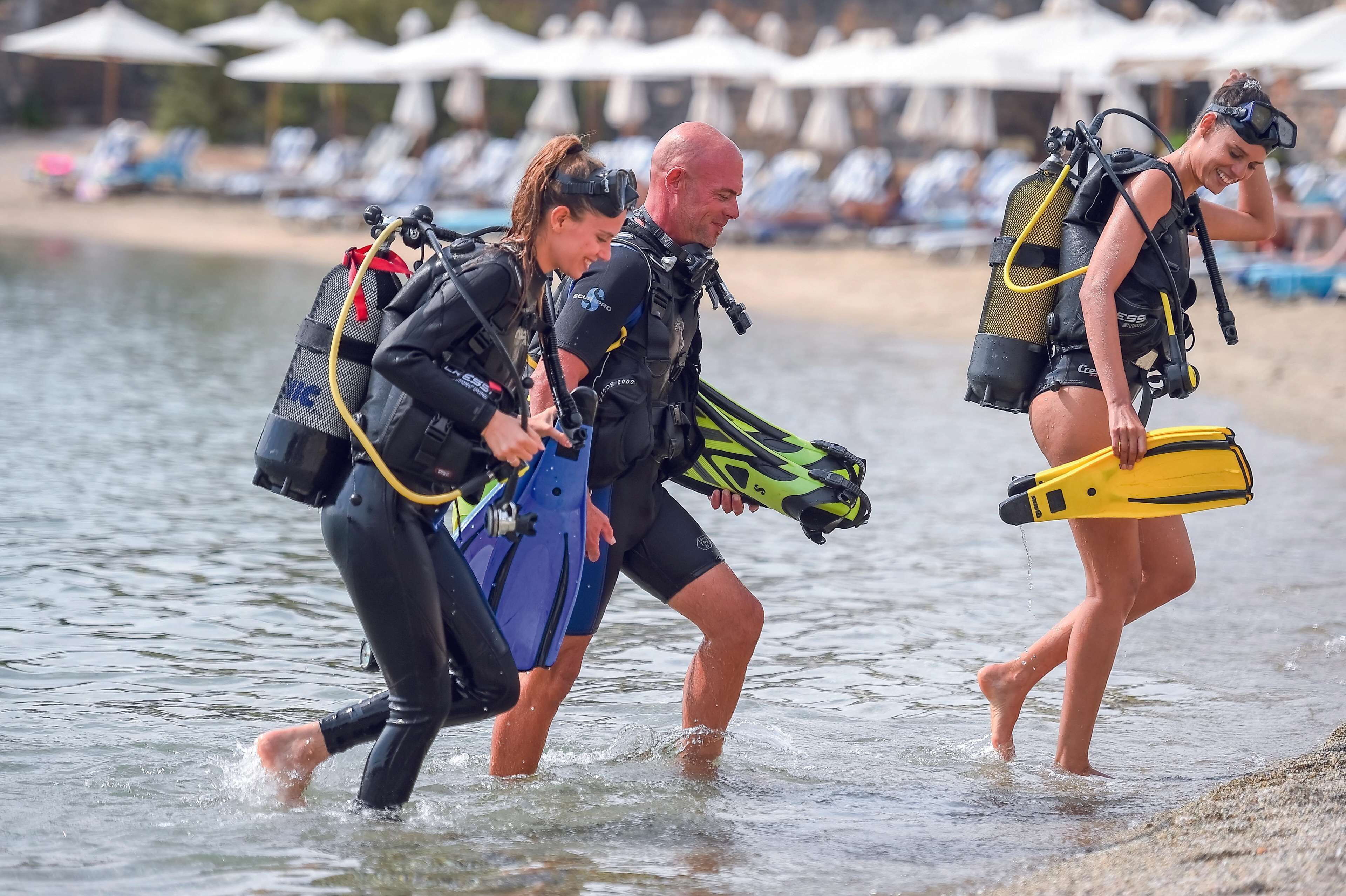 Tres personas con trajes de neopreno y equipo de buceo caminando hacia el agua.