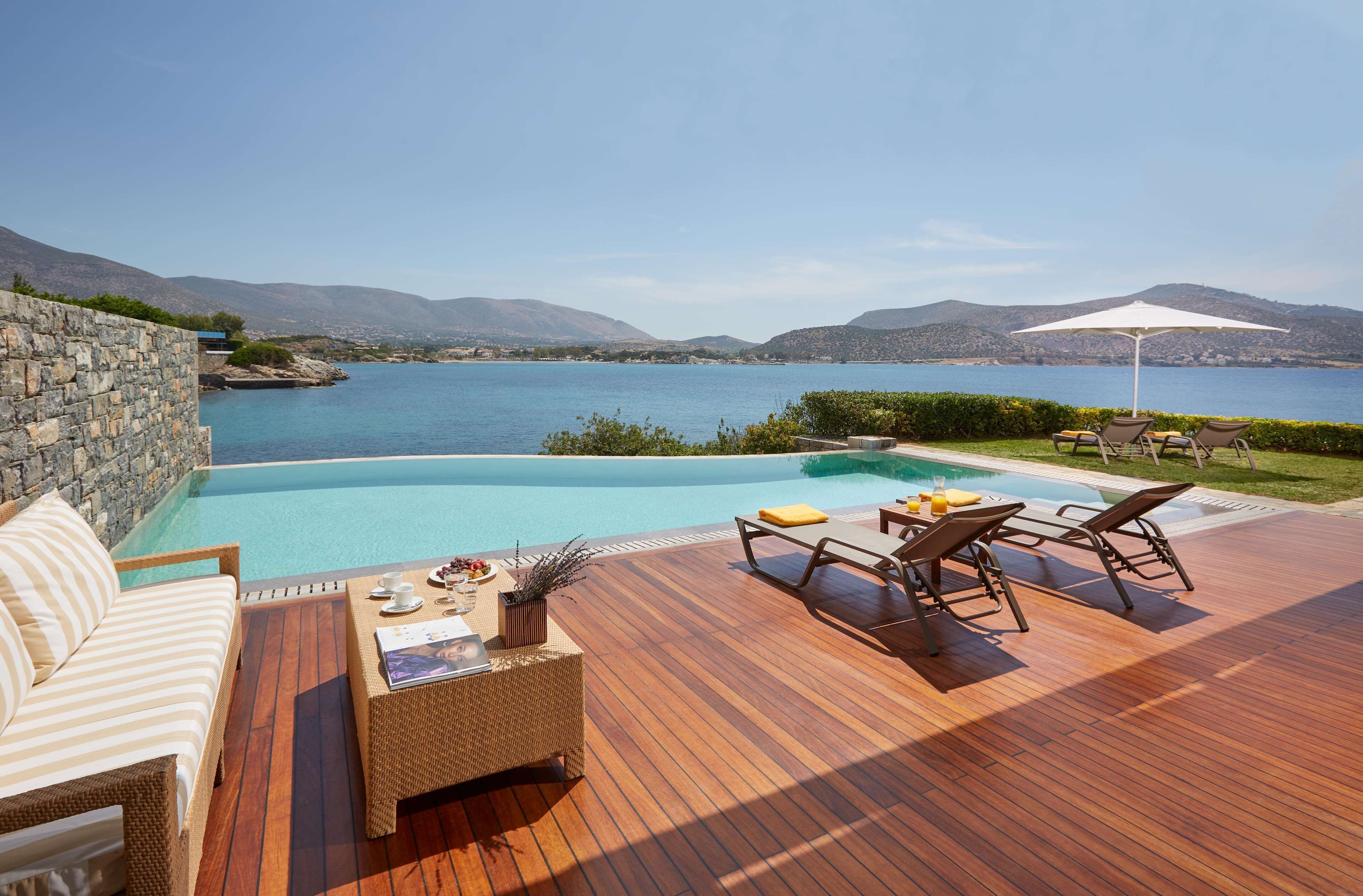 A wooden deck with a pool, lounge chairs, and a sofa overlooking a body of water with mountains in the background.