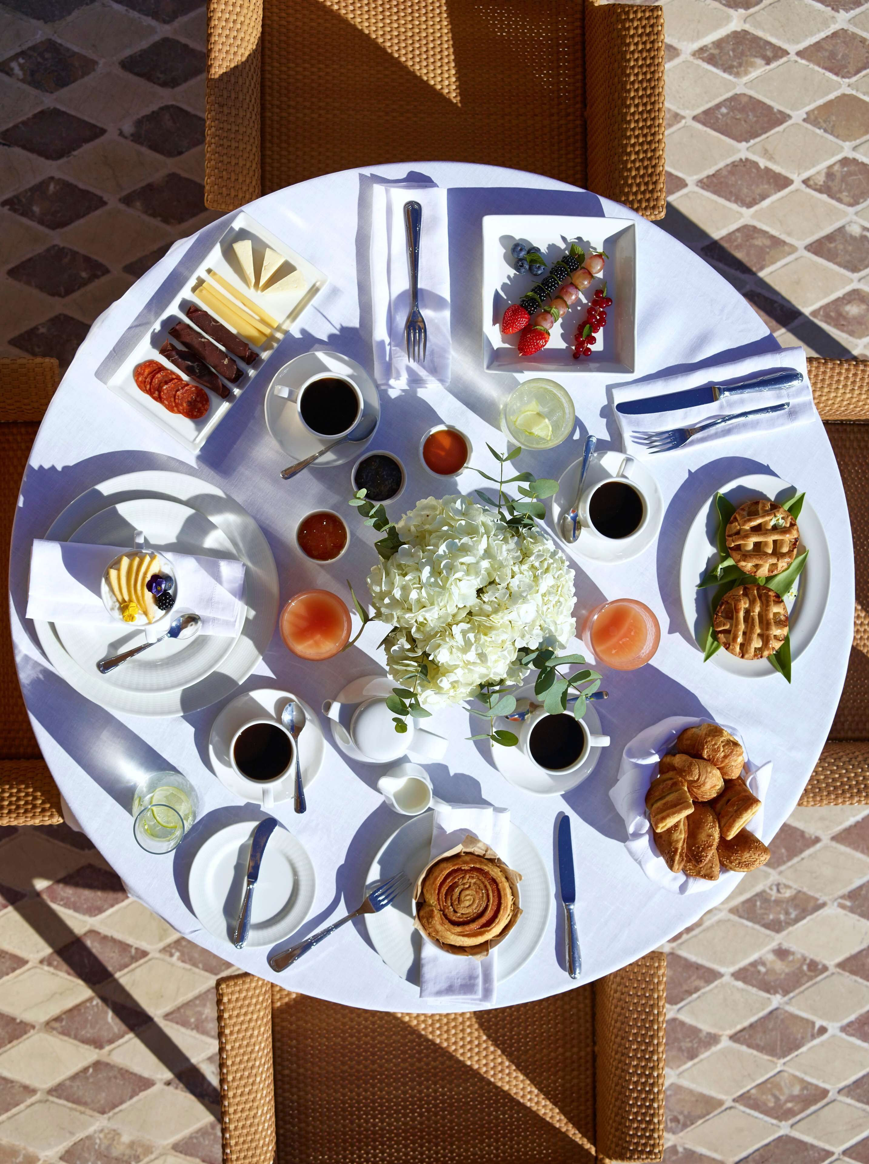 A round table with a variety of food items and drinks, including pastries, fruits, and condiments, arranged on a white tablecloth.