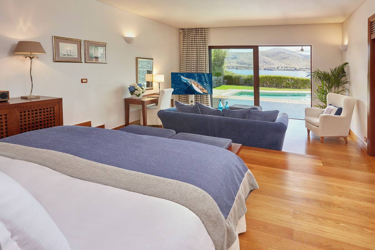 Ein Hotelzimmer mit einem großen Bett, einem Schlafsofa, einem Schreibtisch mit Lampe sowie Blick auf einen Pool und Berge.