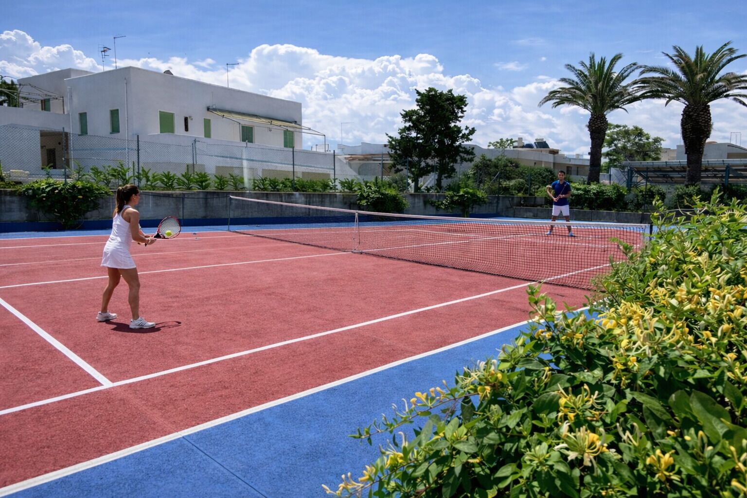 Ein Tennisplatz mit zwei Spielern, umgeben von Grünflächen und Gebäuden.