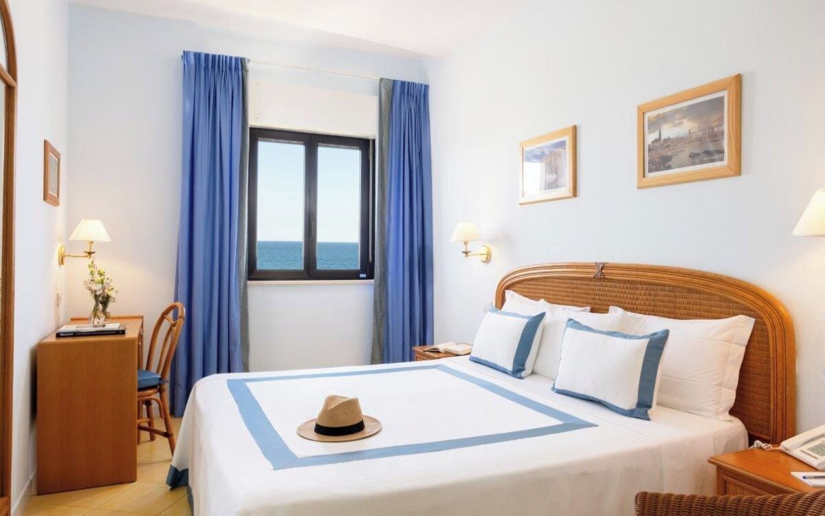 A hotel room with a bed, a desk, a chair, a window with curtains, and two wall-mounted lamps.
