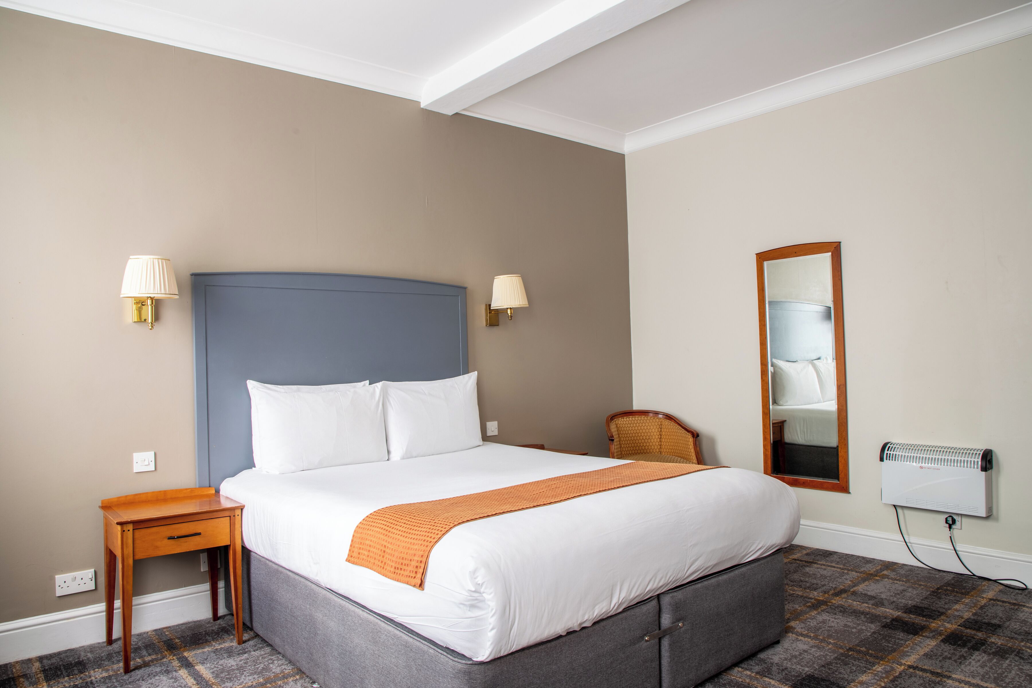 A hotel room with a large bed, a bedside table, a chair, a mirror, and a radiator.