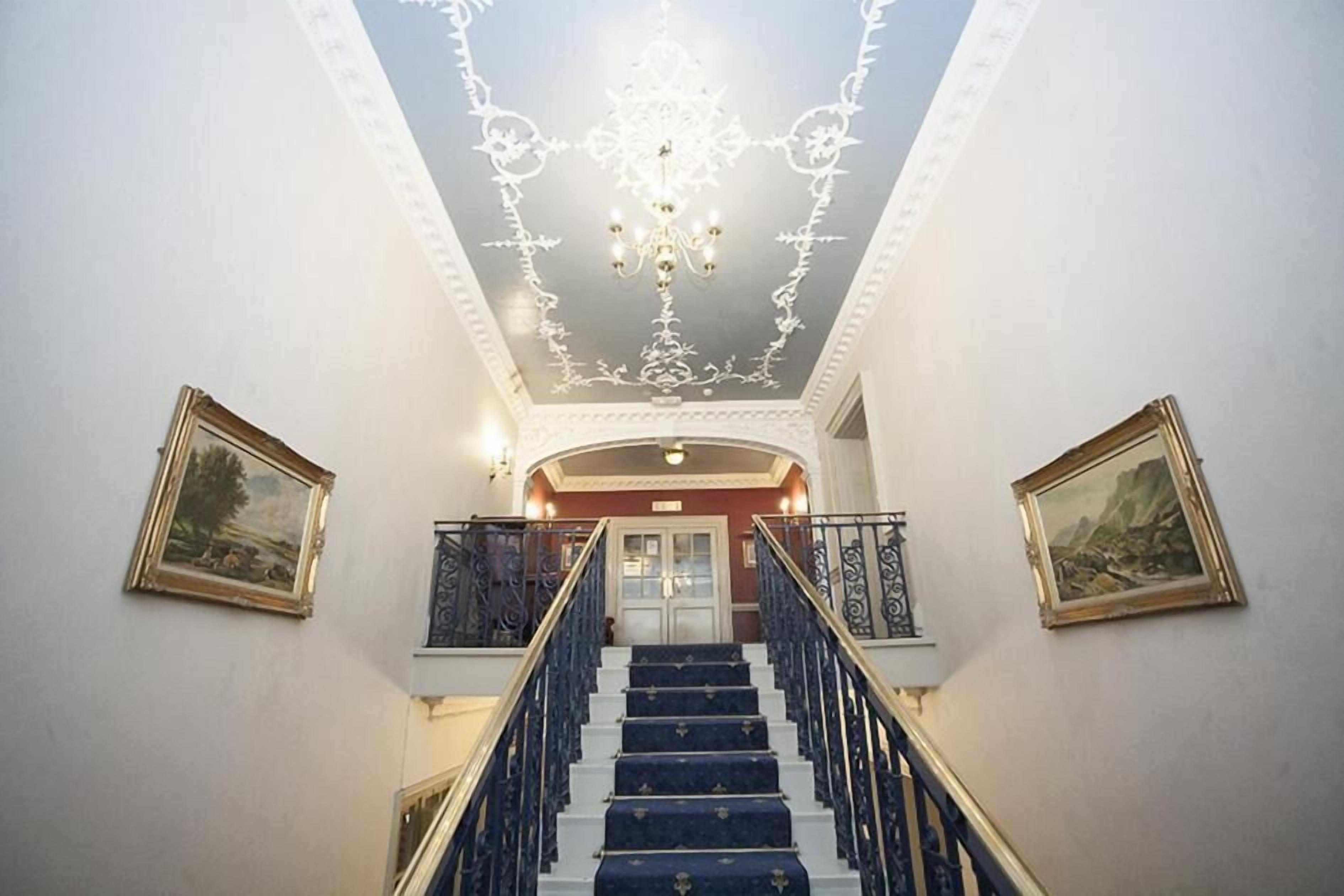 A grand staircase with ornate railings and a chandelier, leading to a door with a red frame.