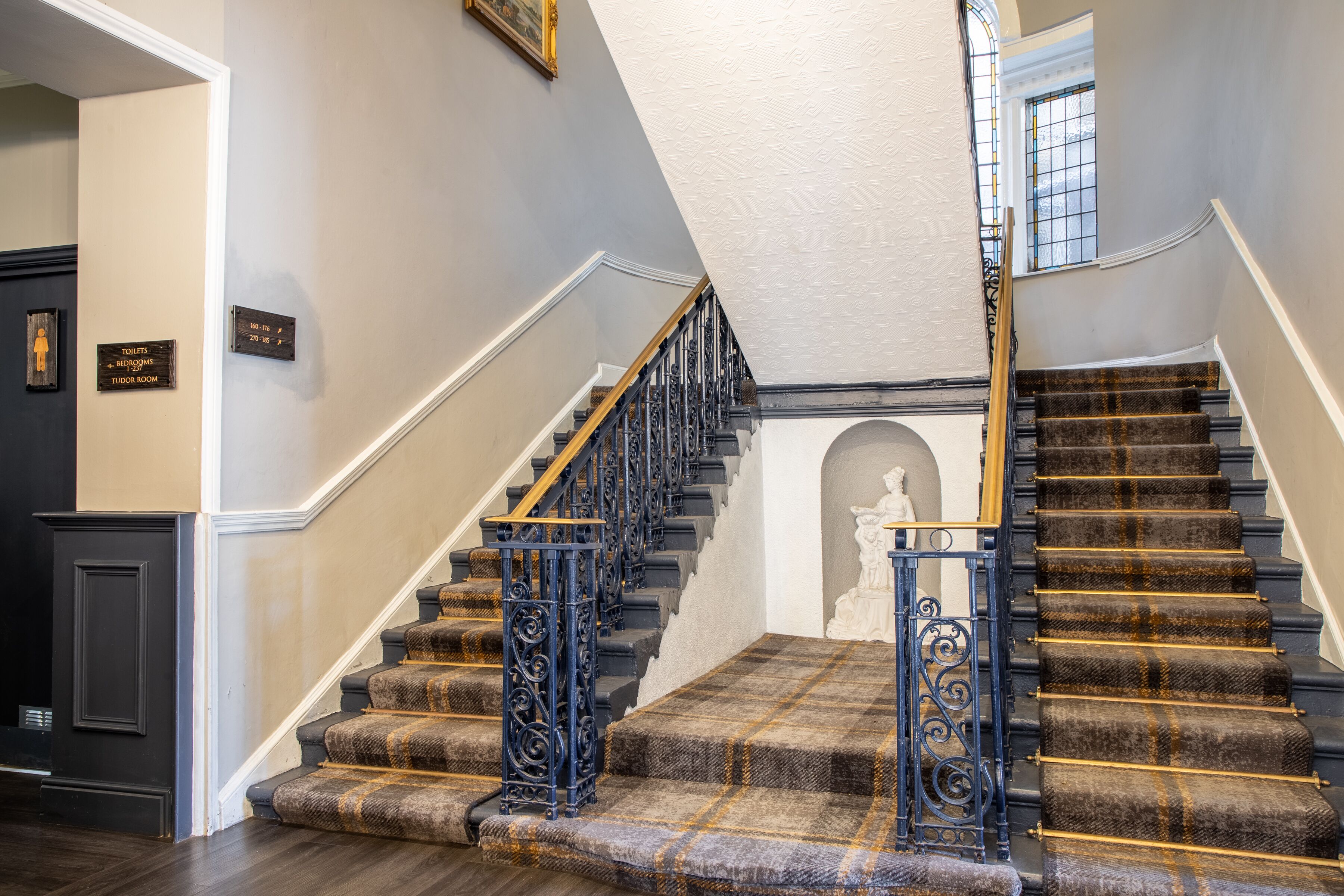 A grand staircase with a checkered carpet and black balusters, leading to a landing with a statue.